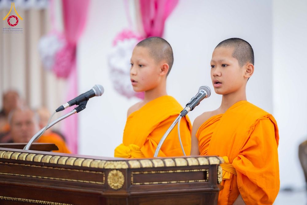 สามเณรสองรูปยืนกล่าวสุนทรพจน์ผ่านไมโครโฟนบนโพเดียมไม้ในงานพิธี