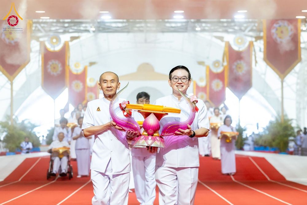 ชายสองคนสวมชุดขาวถือพานพุ่มผ้าไตรจีวรเดินนำขบวนในพิธีทางศาสนา