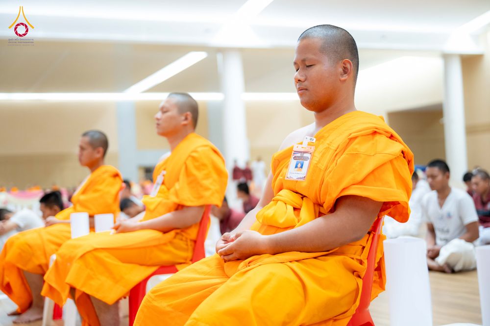 พระภิกษุสงฆ์หลายรูปนั่งสมาธิหลับตาในจีวรสีเหลืองทองภายในห้องโถง