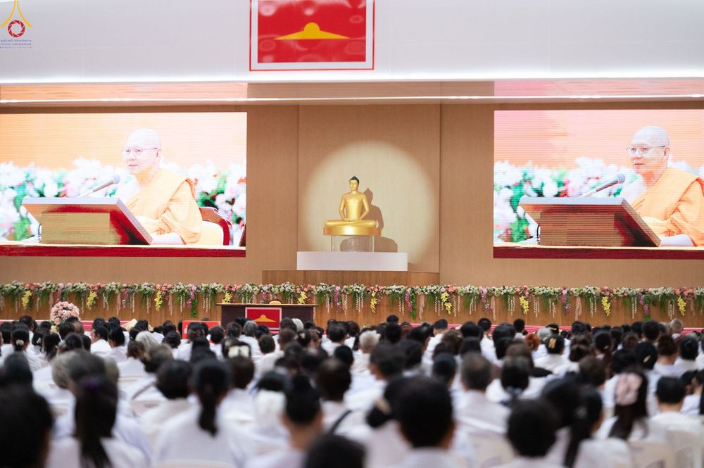 ผู้เข้าร่วมงานจำนวนมากนั่งฟังการบรรยายธรรมภายในหอประชุม โดยมีจอภาพขนาดใหญ่แสดงภาพพระภิกษุสงฆ์และมีพระพุทธรูปอยู่บนเวที