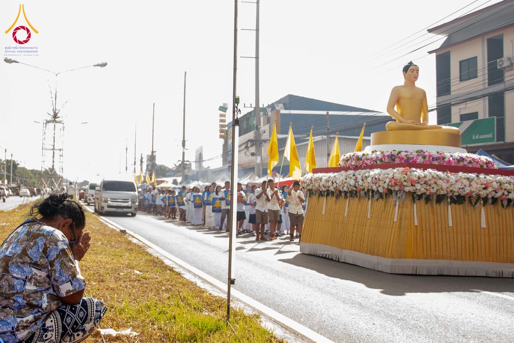 ขบวนรถแห่พระพุทธรูปบนถนนที่มีผู้คนเดินตามและหญิงสูงวัยนั่งไหว้ริมทาง