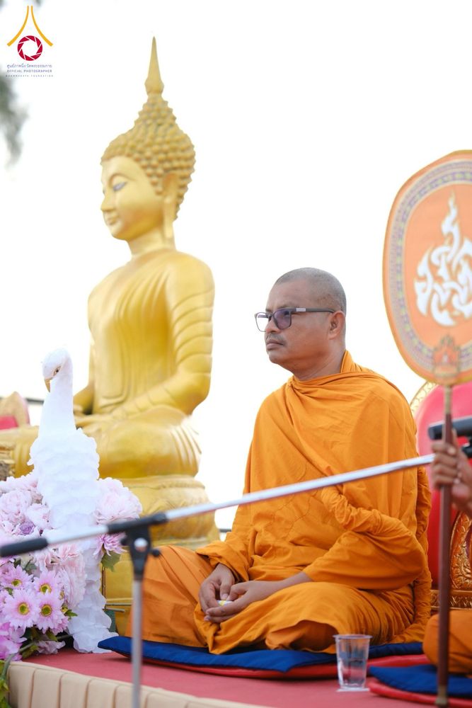 พระภิกษุสงฆ์สวมจีวรสีส้มและแว่นตานั่งอยู่หน้าพระพุทธรูปสีทองในพิธีทางศาสนา