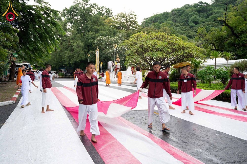 กลุ่มนักเรียนนายสิบตำรวจกำลังช่วยกันปูผ้าสีขาวและสีชมพูบนพื้นถนนเพื่อเตรียมพื้นที่พิธีตักบาตร