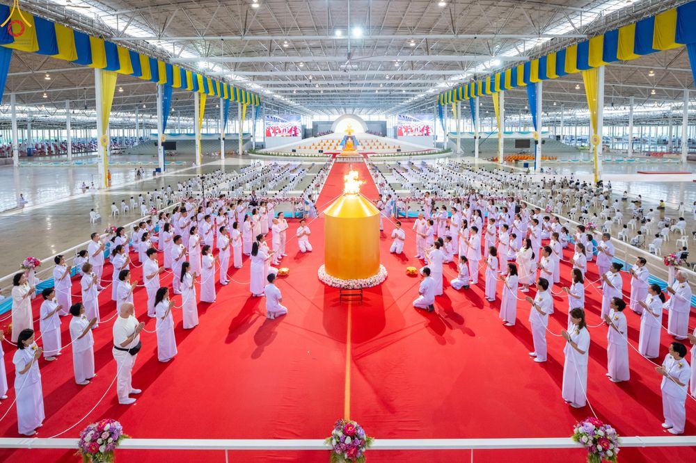 กลุ่มคนสวมชุดสีขาวกำลังประกอบพิธีรอบโคมเทียนสีทองขนาดใหญ่บนพรมสีแดงภายในอาคาร