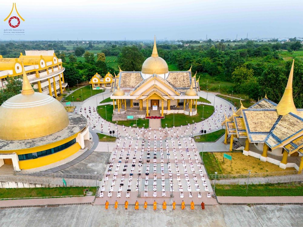 กลุ่มผู้ปฏิบัติธรรมชุดขาวนั่งสมาธิเป็นแถวหน้าอาคารทรงไทยสีทองท่ามกลางพื้นที่สีเขียว
