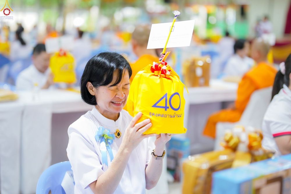 หญิงไทยสวมชุดสีขาวถือถุงผ้าสีเหลืองสกรีนเลข 40 ด้วยสีหน้ายิ้มแย้มมีความสุขภายในงานบุญ