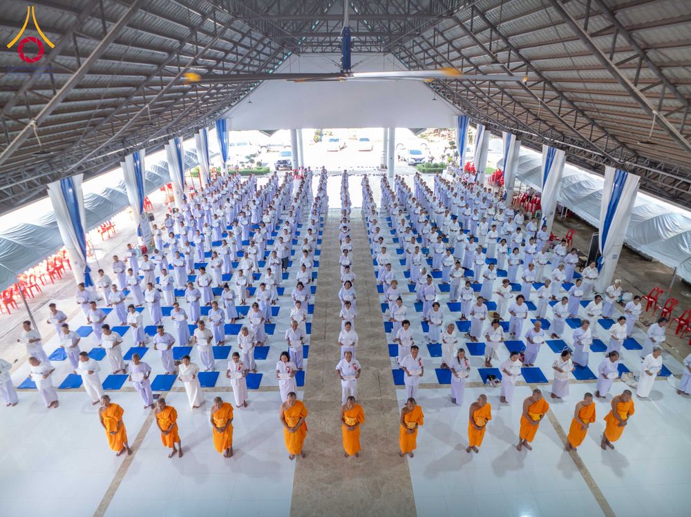 กลุ่มผู้เข้าร่วมโครงการอบรมในชุดสีขาวจำนวนมากยืนเรียงแถวภายในอาคารปฏิบัติธรรม โดยมีพระภิกษุในชุดจีวรสีส้มยืนนำอยู่ด้านหน้า