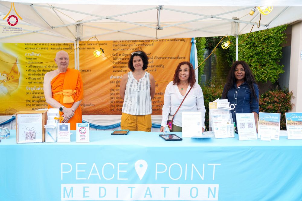 พระภิกษุและสตรีสามท่านยืนเรียงแถวหน้าโต๊ะลงทะเบียนที่มีป้าย Peace Point Meditation ภายใต้เต็นท์สีขาว