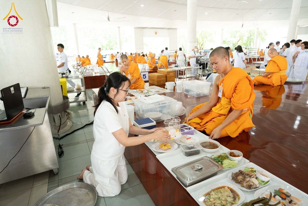 หญิงชุดขาวกำลังประเคนอาหารแด่พระภิกษุสงฆ์ภายในหอฉันที่มีผู้คนร่วมงานจำนวนมาก