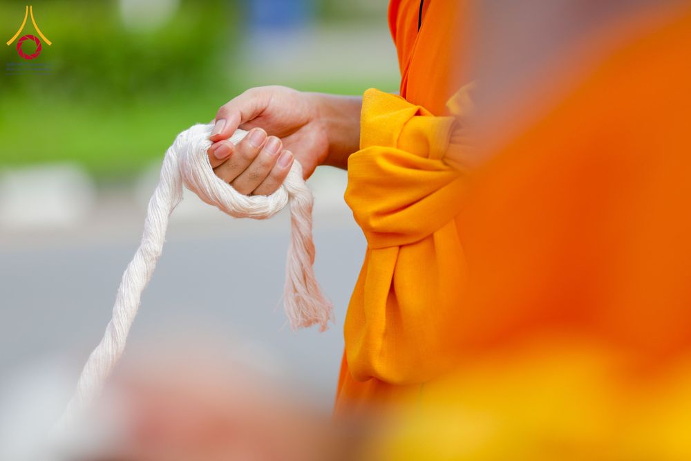 มือพระภิกษุสวมจีวรสีส้มกำลังถือสายสิญจน์สีขาวในพิธีกรรม