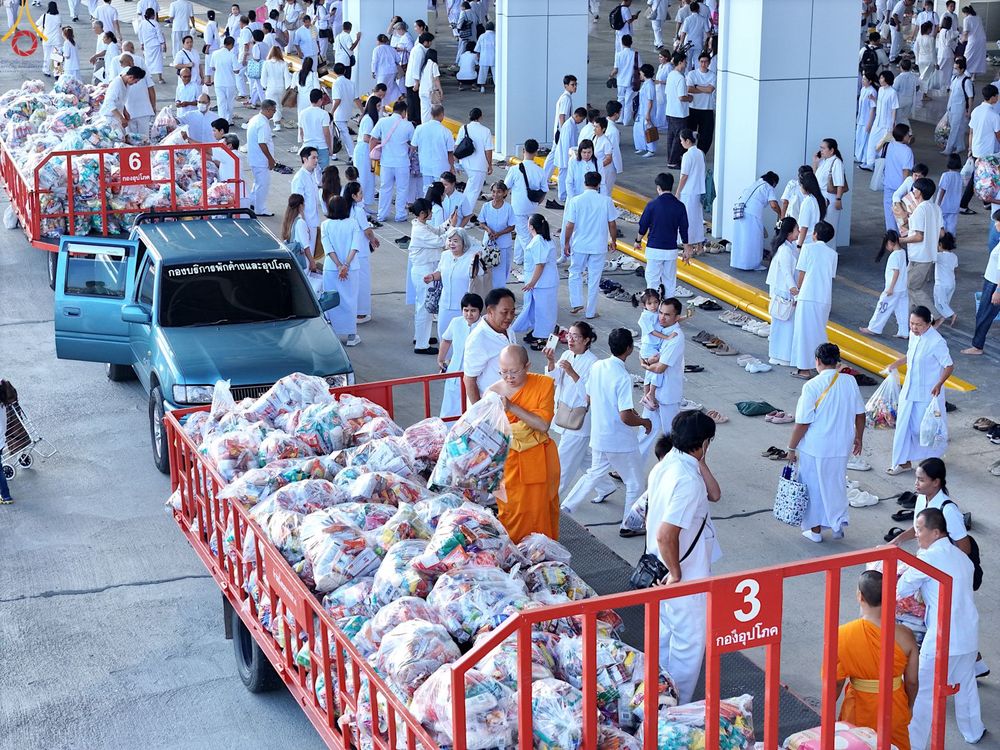 พุทธศาสนิกชนสวมชุดขาวจำนวนมากกำลังร่วมพิธีตักบาตรข้าวสารอาหารแห้ง โดยมีรถกระบะบรรทุกถุงอาหารแห้งจอดอยู่ภายในบริเวณวัด