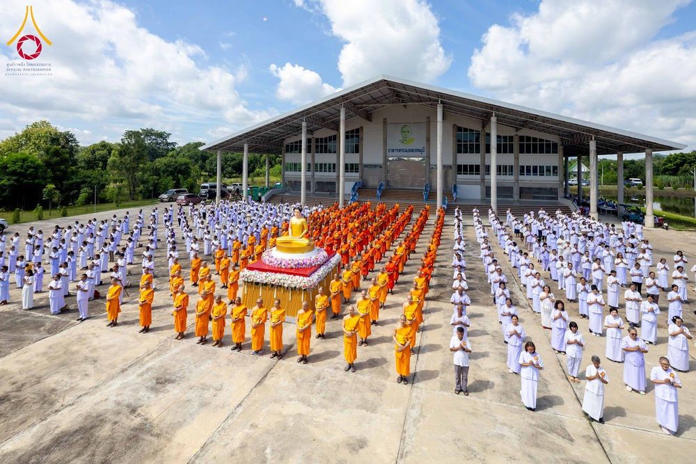 พระภิกษุสงฆ์และสาธุชนชุดขาวจำนวนมากร่วมพิธีกรรมกลางแจ้งหน้าอาคาร โดยมีพระพุทธรูปประดิษฐานอยู่บนแท่นประดับดอกไม้