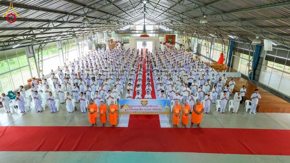 กลุ่มผู้เข้าร่วมโครงการอบรมในชุดสีขาวนั่งเรียงแถวภายในอาคารโดยมีพระภิกษุสงฆ์ยืนอยู่ด้านหน้า