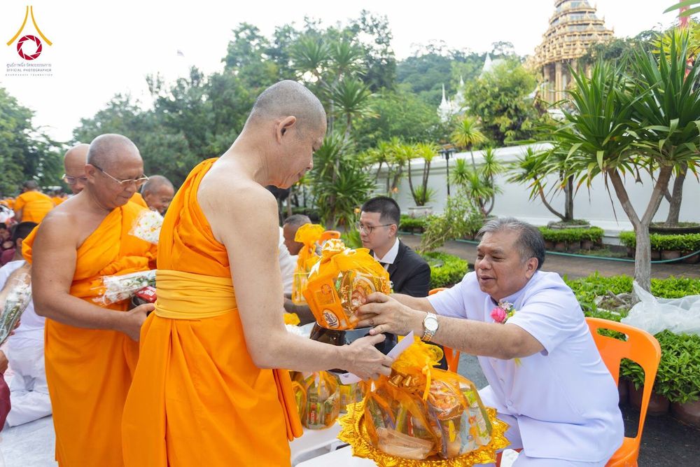 ชายสวมชุดขาวกำลังประเคนของใส่บาตรให้สามเณรในพิธีตักบาตร