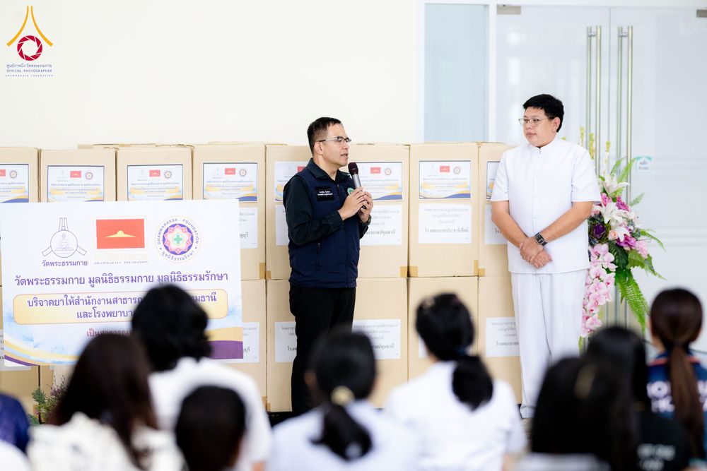 ผู้แทนกล่าวรายงานในพิธีมอบยาและเวชภัณฑ์ โดยมีกล่องพัสดุจำนวนมากวางเรียงรายอยู่ด้านหลัง