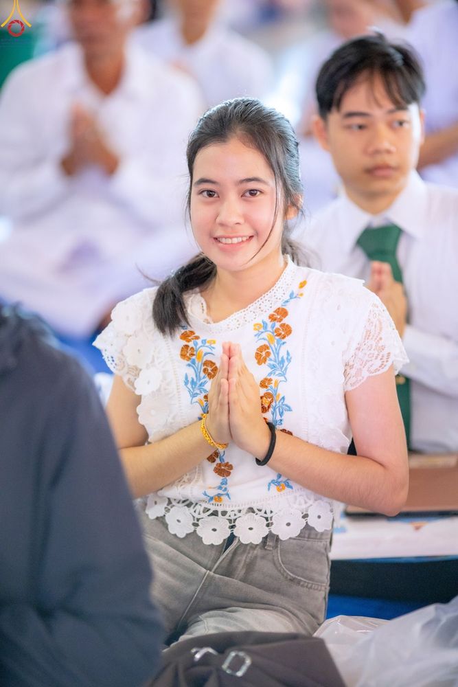 หญิงสาวสวมเสื้อสีขาวปักลายดอกไม้กำลังพนมมือไหว้ด้วยรอยยิ้ม