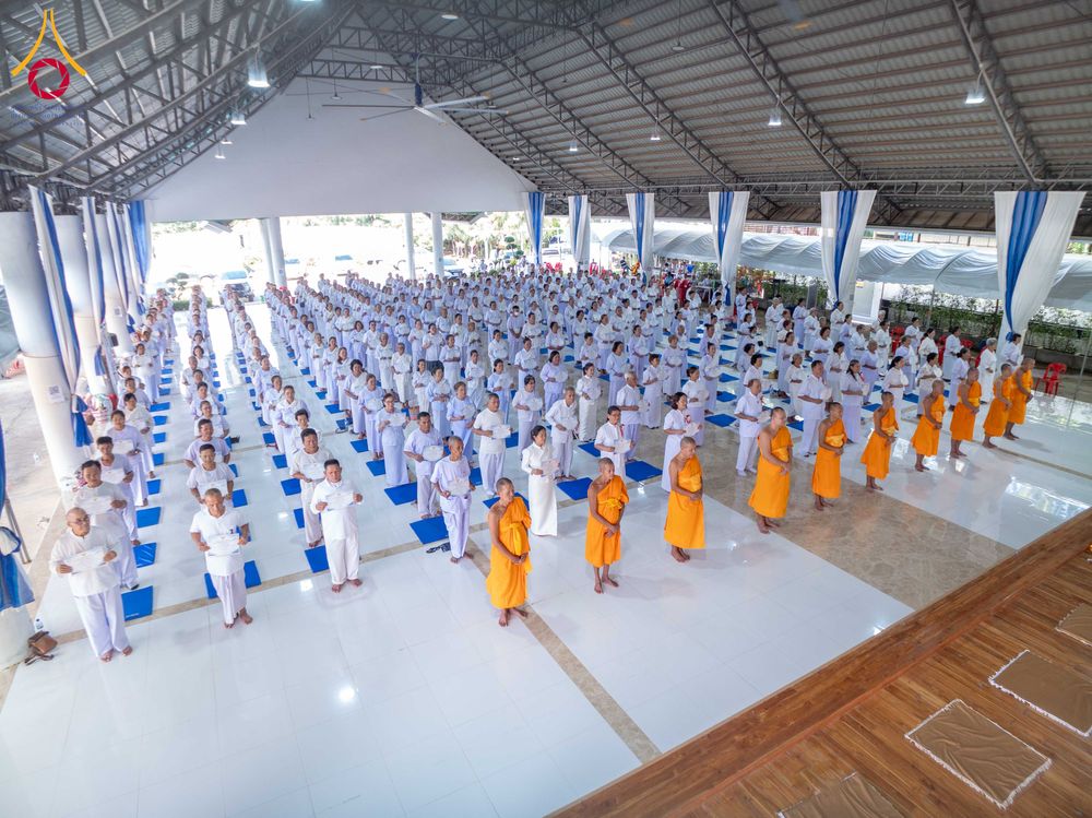 กลุ่มผู้เข้าร่วมโครงการอบรมสวมชุดสีขาวและพระภิกษุสวมจีวรสีเหลืองยืนเรียงแถวทำกิจกรรมภายในอาคาร