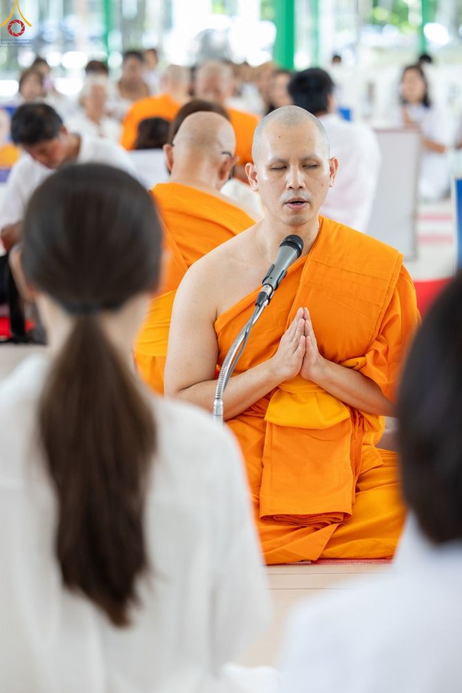 พระภิกษุสงฆ์นั่งพนมมือถือไมโครโฟนนำสวดมนต์ท่ามกลางผู้เข้าร่วมพิธีในชุดสีขาว