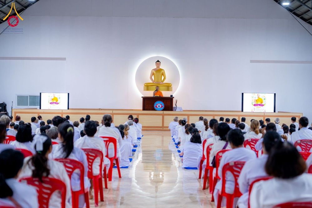 กลุ่มผู้เข้าร่วมอบรมสวมชุดขาวนั่งฟังธรรมจากพระภิกษุบนเวทีภายในอาคาร