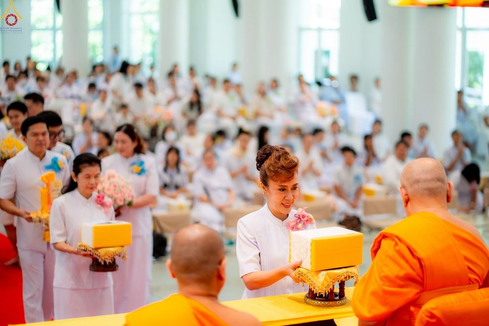 หญิงไทยในชุดขาวกำลังประคองพานถวายสังฆทานแด่พระภิกษุสงฆ์ท่ามกลางพุทธศาสนิกชนที่มาร่วมงานบุญ