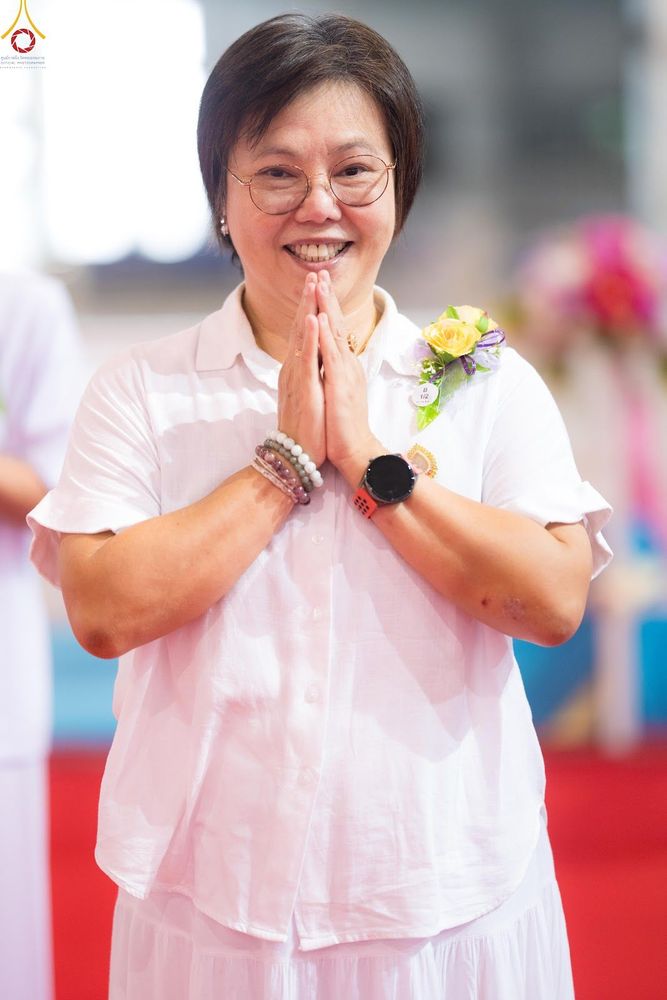 หญิงวัยกลางคนสวมชุดสีขาวพนมมือไหว้ด้วยรอยยิ้ม
