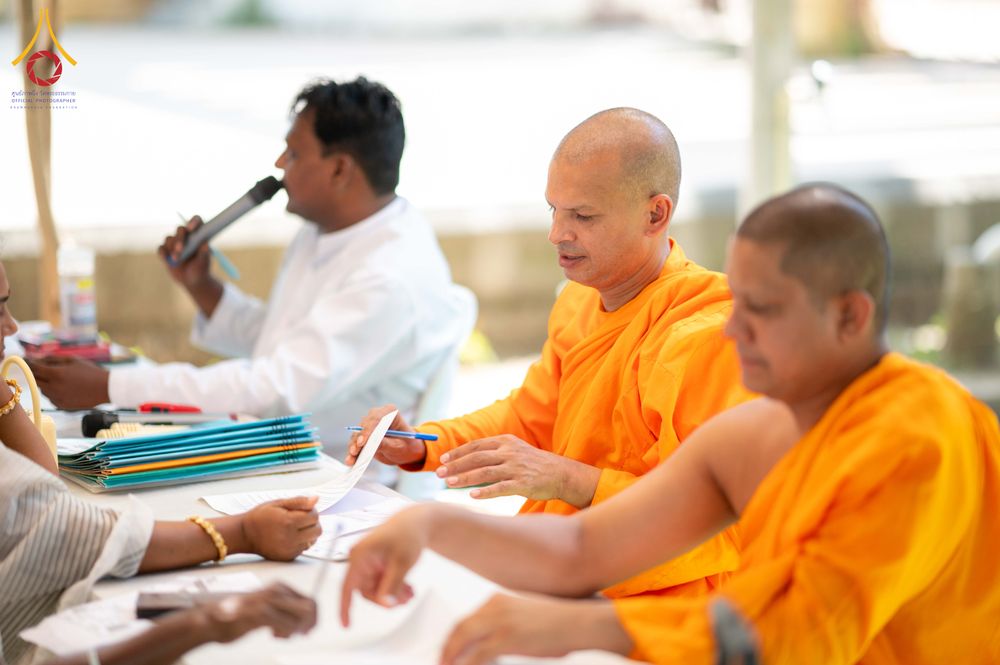 พระภิกษุสงฆ์ในจีวรสีส้มและเจ้าหน้าที่กำลังนั่งปฏิบัติงานลงทะเบียนเอกสารให้กับผู้เข้าร่วมโครงการ