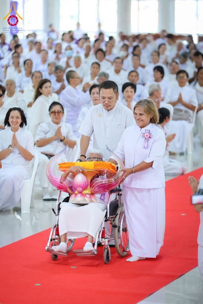 กลุ่มคนในชุดขาวกำลังเข็นรถเข็นที่มีพานพุ่มวางผ้าไตรสีเหลืองทองบนพรมสีแดงภายในอาคาร