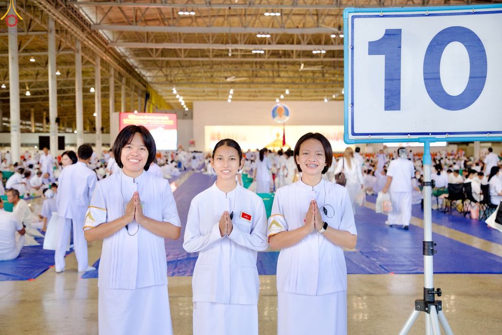 หญิงสาวสามคนสวมชุดสีขาวพนมมือยิ้มแย้มในโถงกว้างที่มีผู้คนร่วมกิจกรรมทางศาสนาและมีป้ายหมายเลข 10