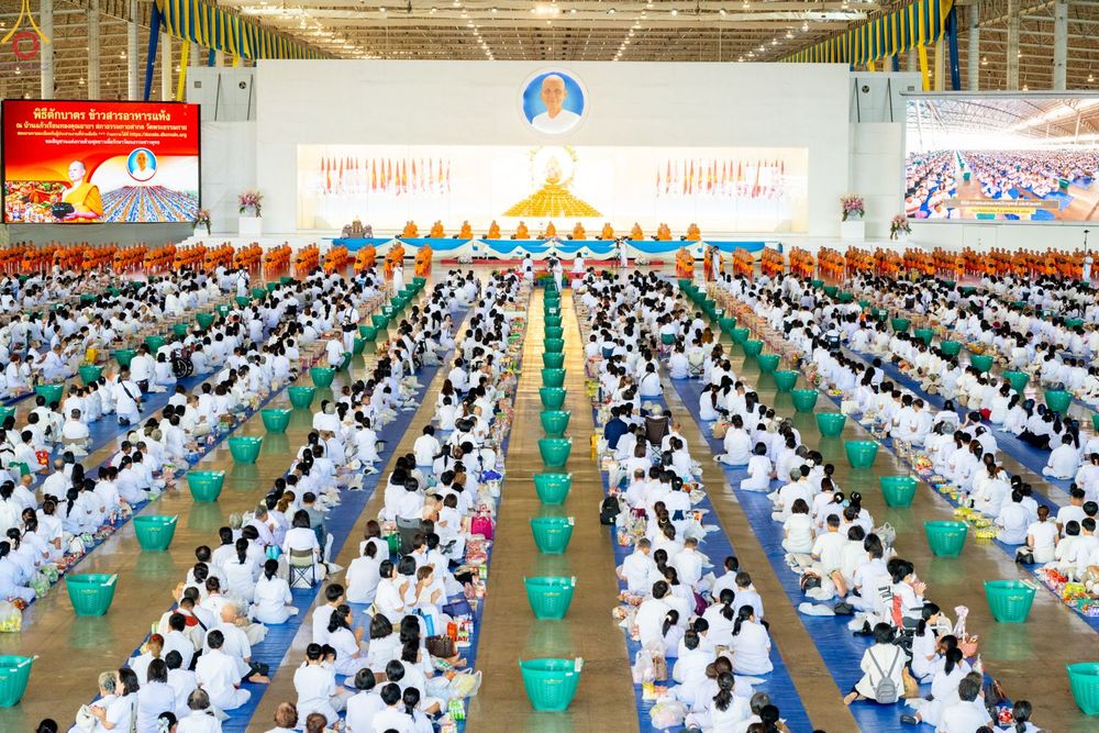 พุทธศาสนิกชนสวมชุดขาวนั่งเรียงแถวร่วมพิธีตักบาตรภายในอาคารขนาดใหญ่ที่มีพระภิกษุสงฆ์นั่งอยู่ด้านหน้า