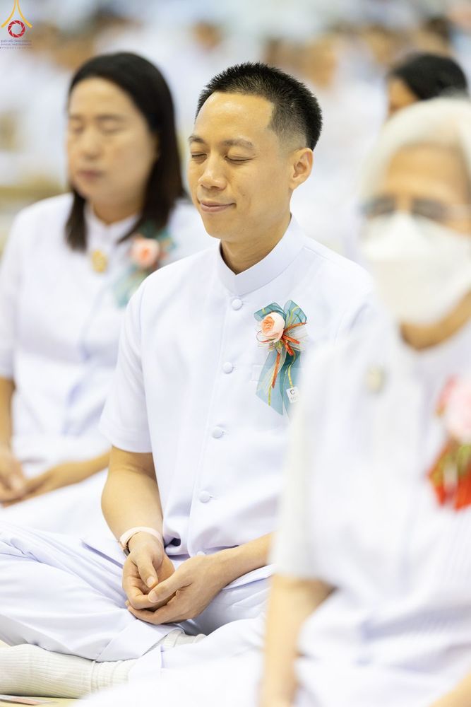 ชายหนุ่มสวมชุดสีขาวติดเข็มกลัดดอกไม้กำลังนั่งหลับตาทำสมาธิร่วมกับผู้อื่นในพิธีทางศาสนา