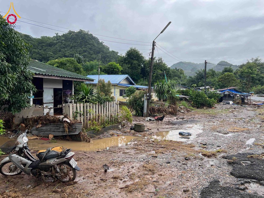 สภาพถนนและบ้านเรือนที่มีคราบโคลนและเศษดินหลังเหตุการณ์น้ำท่วม มีรถจักรยานยนต์จอดอยู่ด้านหน้า