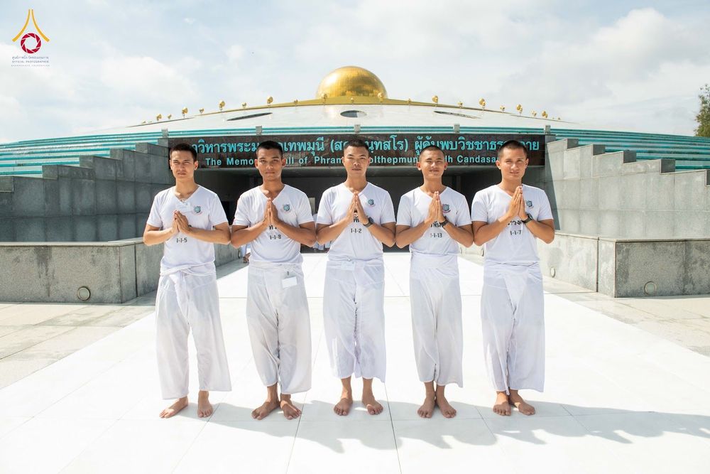 นักเรียนนายสิบตำรวจ 5 นายสวมชุดสีขาวพนมมือยืนเรียงแถวหน้าอาคารสภาธรรมกายสากล