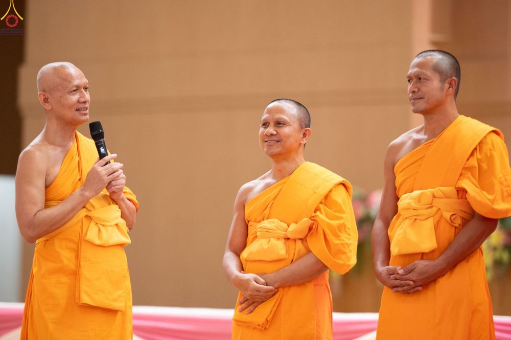 พระภิกษุสงฆ์สามรูปในจีวรสีเหลืองทองยืนอยู่บนเวที รูปซ้ายสุดถือไมโครโฟนพูดคุย อีกสองรูปยืนฟังด้วยความสงบ