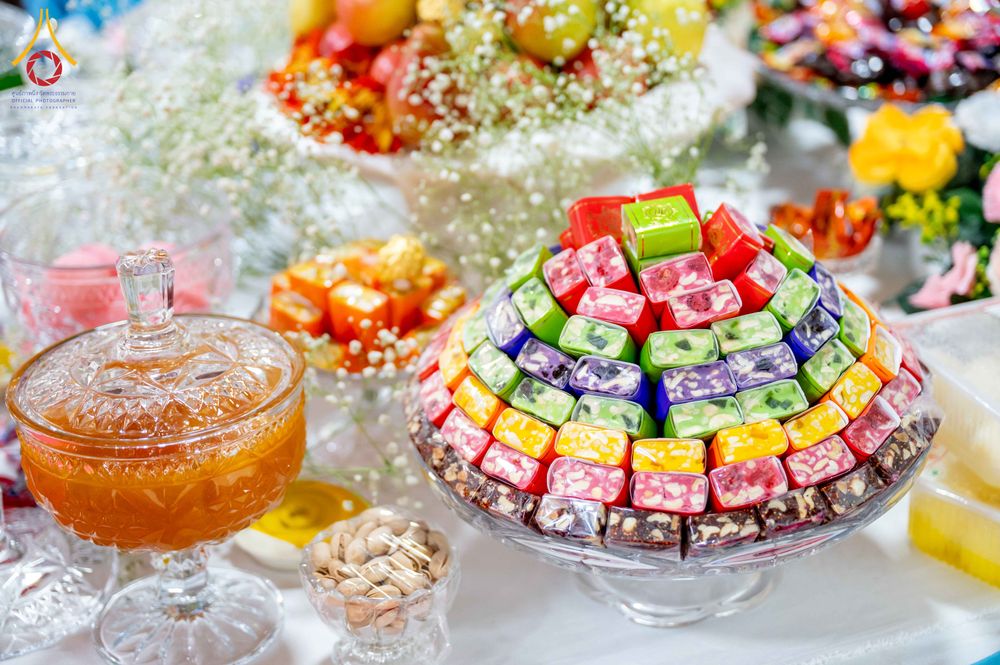 ขนมหวานหลากสีสันจัดเรียงเป็นรูปพีระมิดบนจานแก้ว วางเคียงข้างโถแก้วบรรจุเครื่องดื่มและถั่วบนโต๊ะพิธี