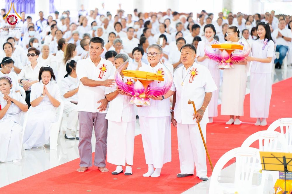 กลุ่มพุทธศาสนิกชนในชุดขาวถือพานพุ่มผ้าไตรยืนบนพรมแดงในพิธีทางศาสนา