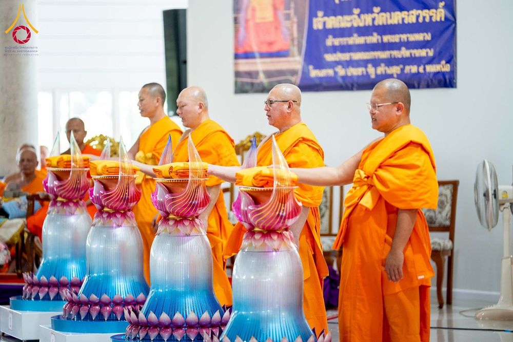 พระภิกษุสงฆ์ในจีวรสีส้มกำลังประกอบพิธีวางผ้าไตรบนพานพุ่มดอกบัวในอาคาร