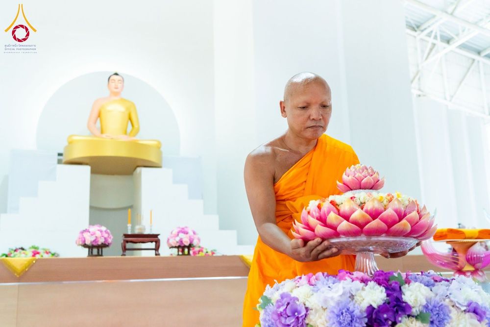 พระภิกษุสงฆ์ถือพานดอกไม้สีชมพูในพิธีทางศาสนา โดยมีพระพุทธรูปสีทองเป็นฉากหลัง