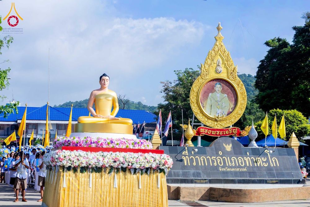 พระพุทธรูปสีทองประดิษฐานบนฐานดอกไม้สด ตั้งอยู่หน้าป้ายที่ว่าการอำเภอท่าตะโกและซุ้มตราสัญลักษณ์