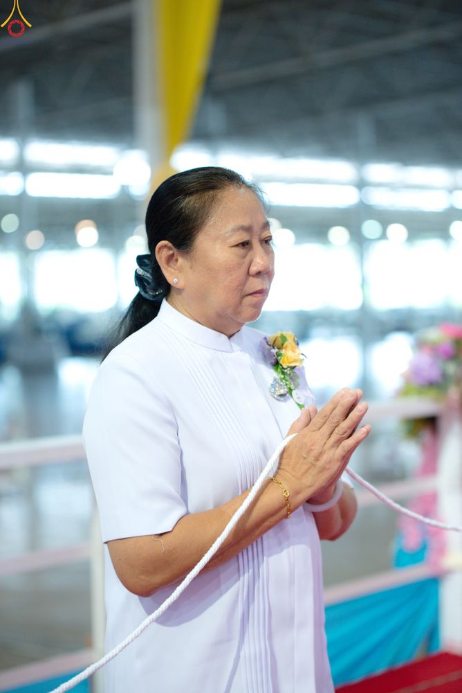 หญิงวัยกลางคนสวมชุดสีขาวพนมมือถือสายสิญจน์ด้วยท่าทางสำรวม