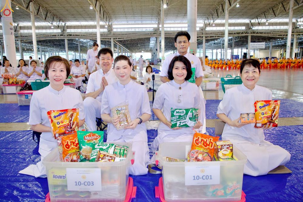 กลุ่มคนสวมชุดสีขาวถือผลิตภัณฑ์อาหารแห้งและเครื่องดื่มนั่งอยู่หน้ากล่องพลาสติกใสภายในอาคาร