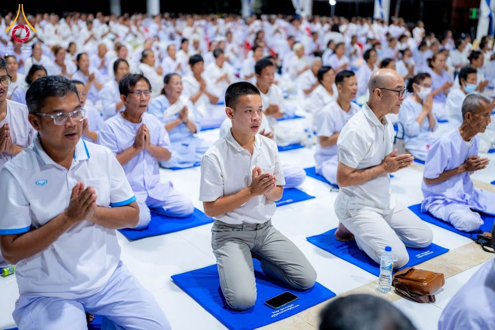 กลุ่มคนสวมชุดสีขาวนั่งพนมมือปฏิบัติธรรมบนเบาะสีน้ำเงินภายในอาคาร