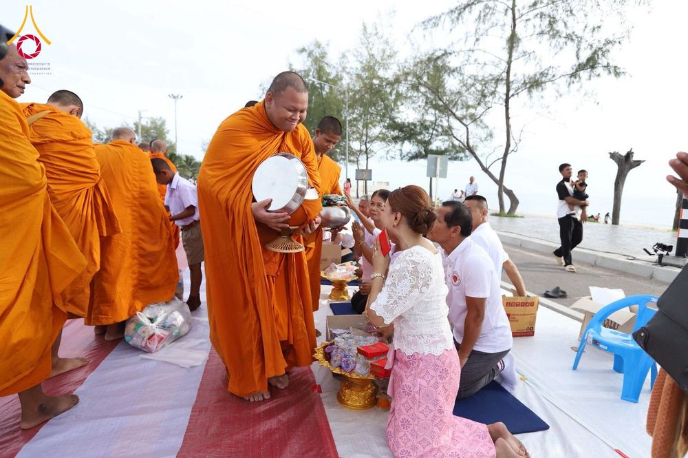 พระสงฆ์ในจีวรสีส้มกำลังรับบิณฑบาตจากพุทธศาสนิกชนที่นั่งคุกเข่าอยู่บนพื้นริมชายหาด