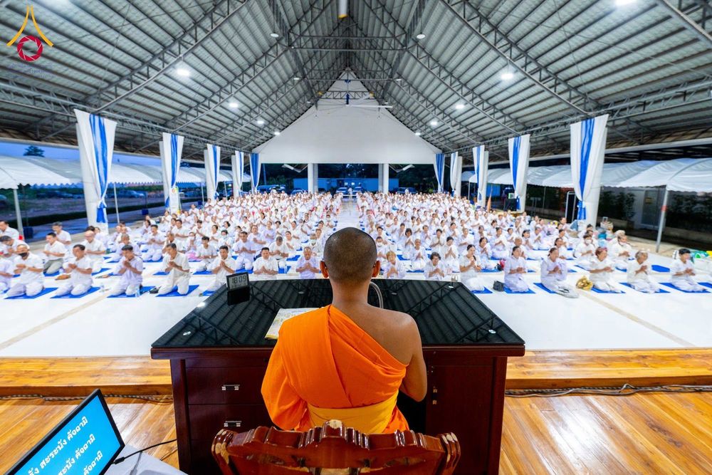 พระภิกษุในชุดจีวรสีส้มกำลังนำพุทธศาสนิกชนจำนวนมากในชุดสีขาวนั่งสมาธิภายในอาคาร