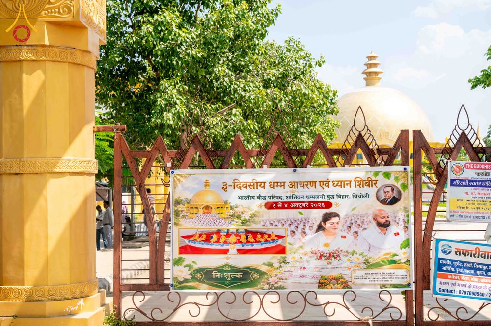 ป้ายไวนิลประชาสัมพันธ์กิจกรรมปฏิบัติธรรมภาษาฮินดีติดตั้งบนรั้วเหล็กหน้าสถานที่ทางศาสนาที่มีเจดีย์สีทองเป็นฉากหลัง