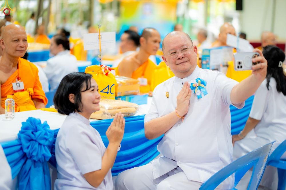 ชายสวมชุดขาวถ่ายภาพเซลฟี่กับหญิงสาวในชุดขาวที่กำลังพนมมือในงานบุญที่มีพระภิกษุสงฆ์ร่วมงาน