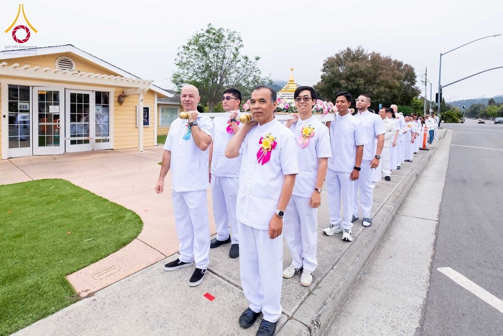 กลุ่มชายสวมชุดสีขาวกำลังช่วยกันแบกบุษบกที่มีพระบรมสารีริกธาตุเดินไปตามทางเท้าหน้าวัด