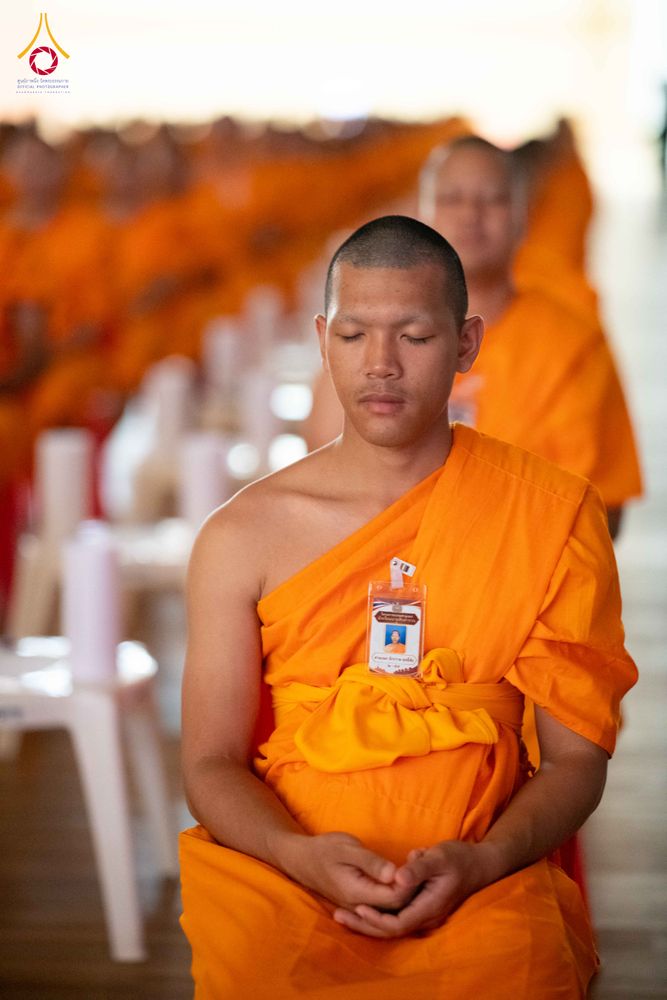 พระภิกษุสงฆ์นั่งหลับตาทำสมาธิในจีวรสีส้ม มีป้ายชื่อติดที่หน้าอก