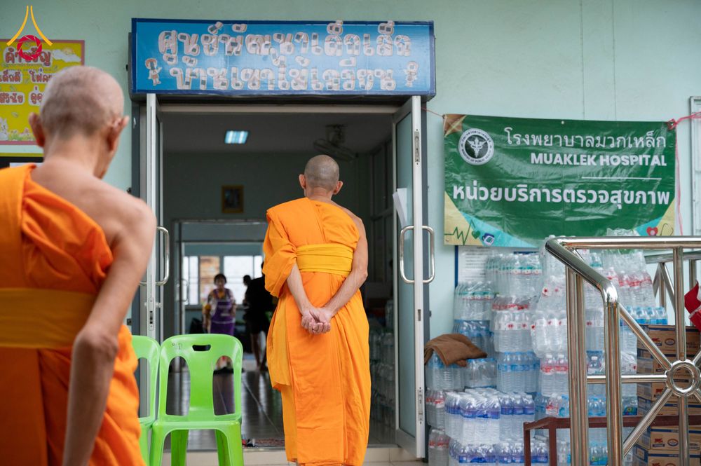 พระสงฆ์ยืนอยู่หน้าอาคารที่มีป้ายโรงพยาบาลมวกเหล็กและมีน้ำดื่มวางเรียงรายอยู่