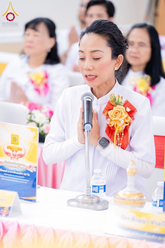 หญิงไทยสวมชุดขาวกำลังกล่าวคำถวายผ้าป่าผ่านไมโครโฟนในพิธีทางศาสนา