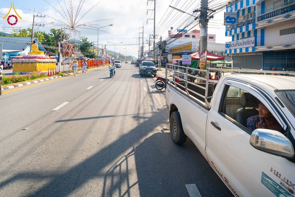 ถนนสายหลักที่มีรถยนต์จอดและวิ่งอยู่ ด้านซ้ายมีขบวนพระภิกษุสงฆ์เดินอยู่ริมทางและแท่นประดิษฐานพระพุทธรูป
